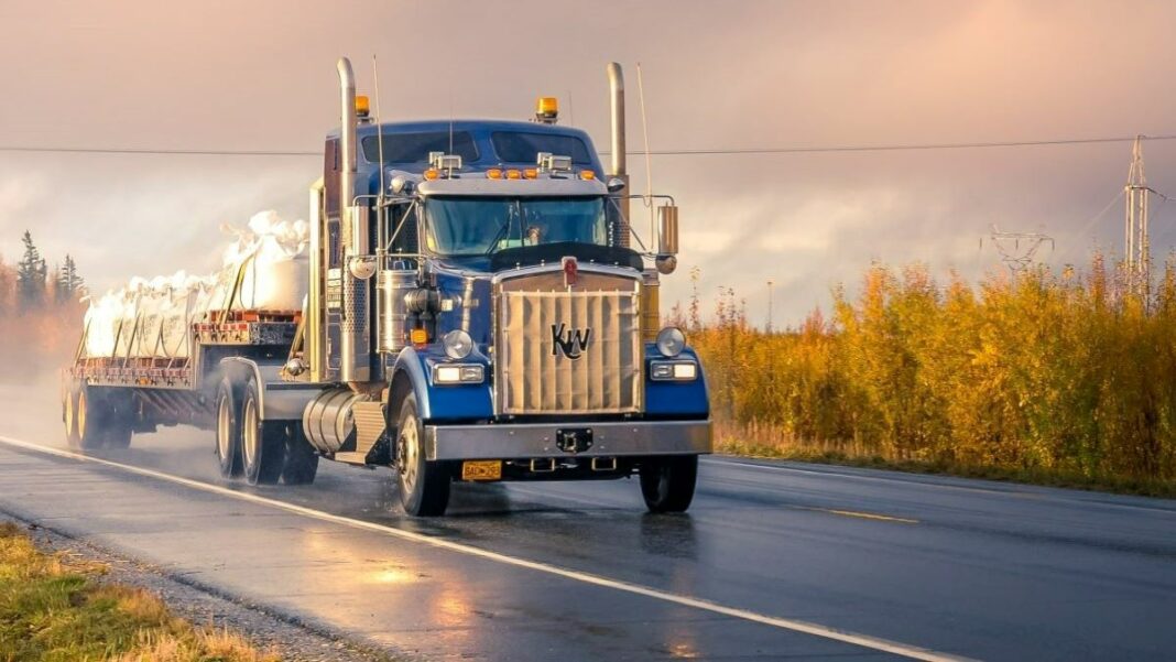 trucking company represented by a shiny blue big rig rolling down a roadway wet with recent rain at sunset