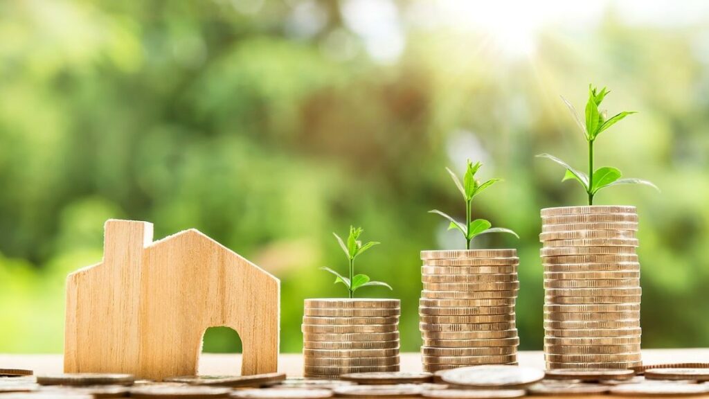 real estate investor represented by a wooden cutout shaped like a house, next to stacks of coins with growing plants set on top of them