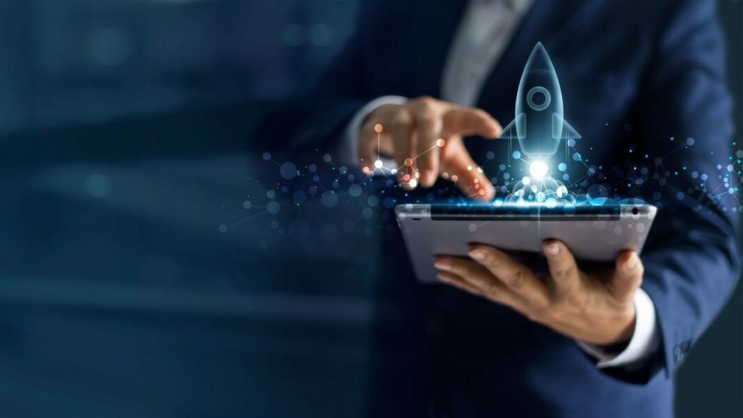 startup accounting represented by a businessman's hands holding a tablet over which a transparent icon of a rocket is launching from the screen with network connection and modern blue background