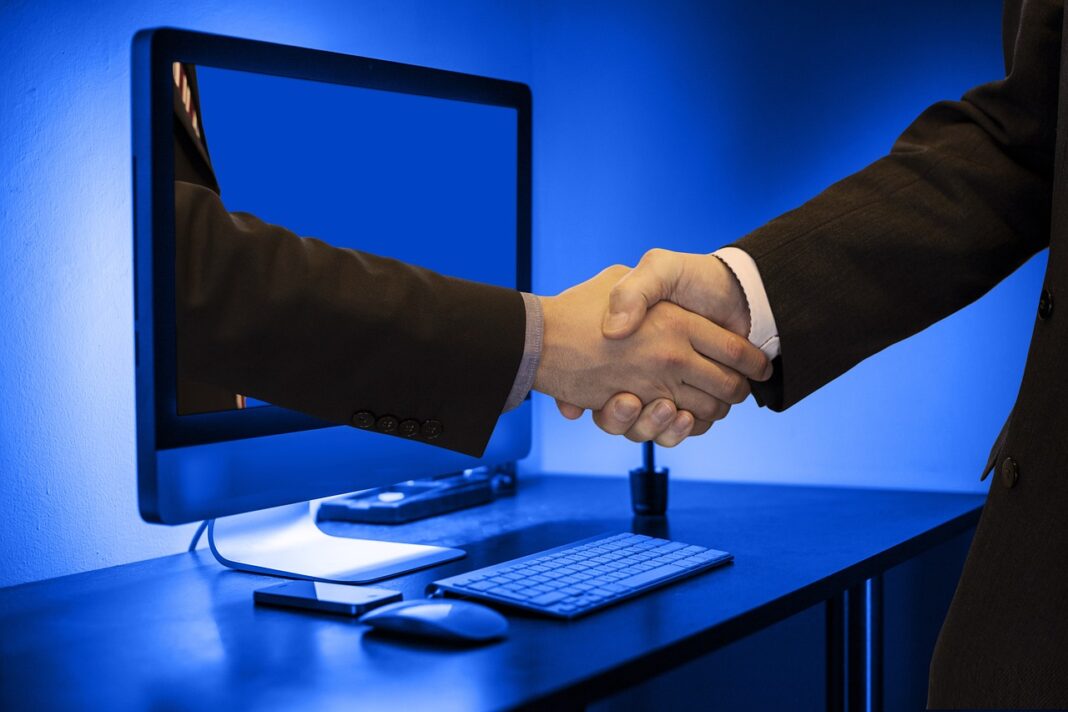 Handshake in front of a computer screen with one hand emerging from the monitor and one from the real world - signifying the online business vs offline business agreement