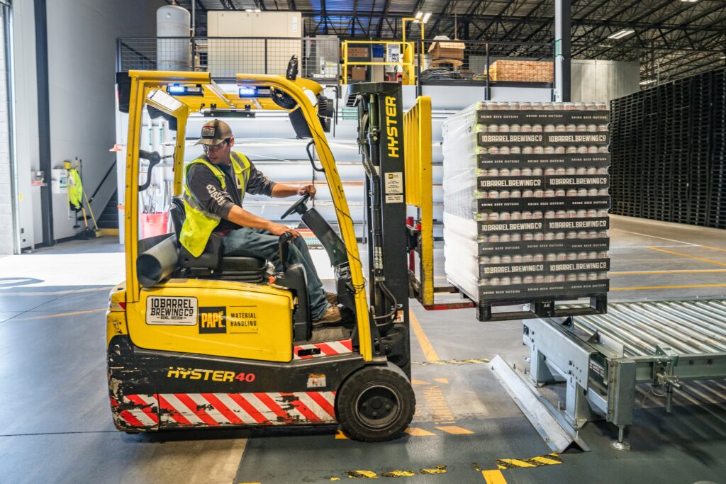 Young, white man in baseball cap and yellow vest operating a forklift in a warehouse