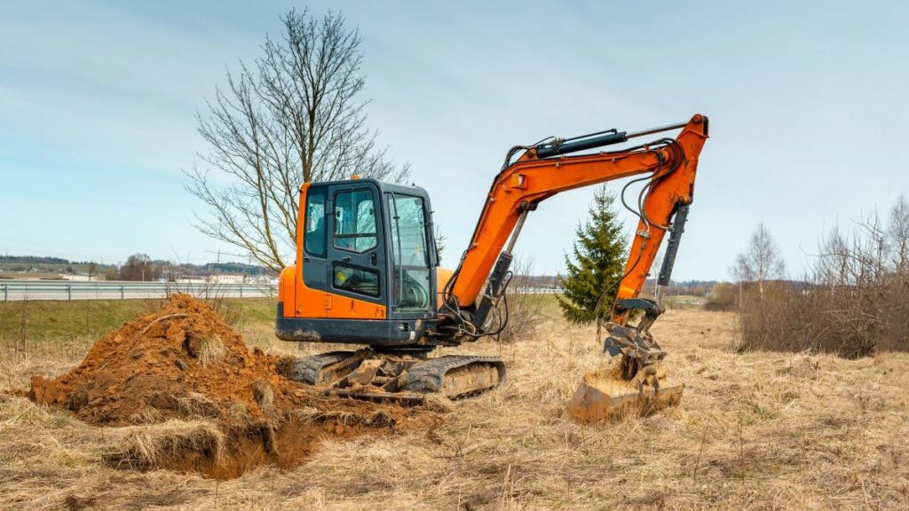 an orange mini excavator digging a trench in an open field, with blue spring or autumn sky