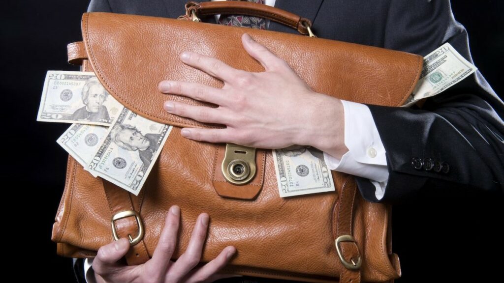 business theft represented by close-up of a man's hands holding a briefcase with money spilling out of it close to his chest