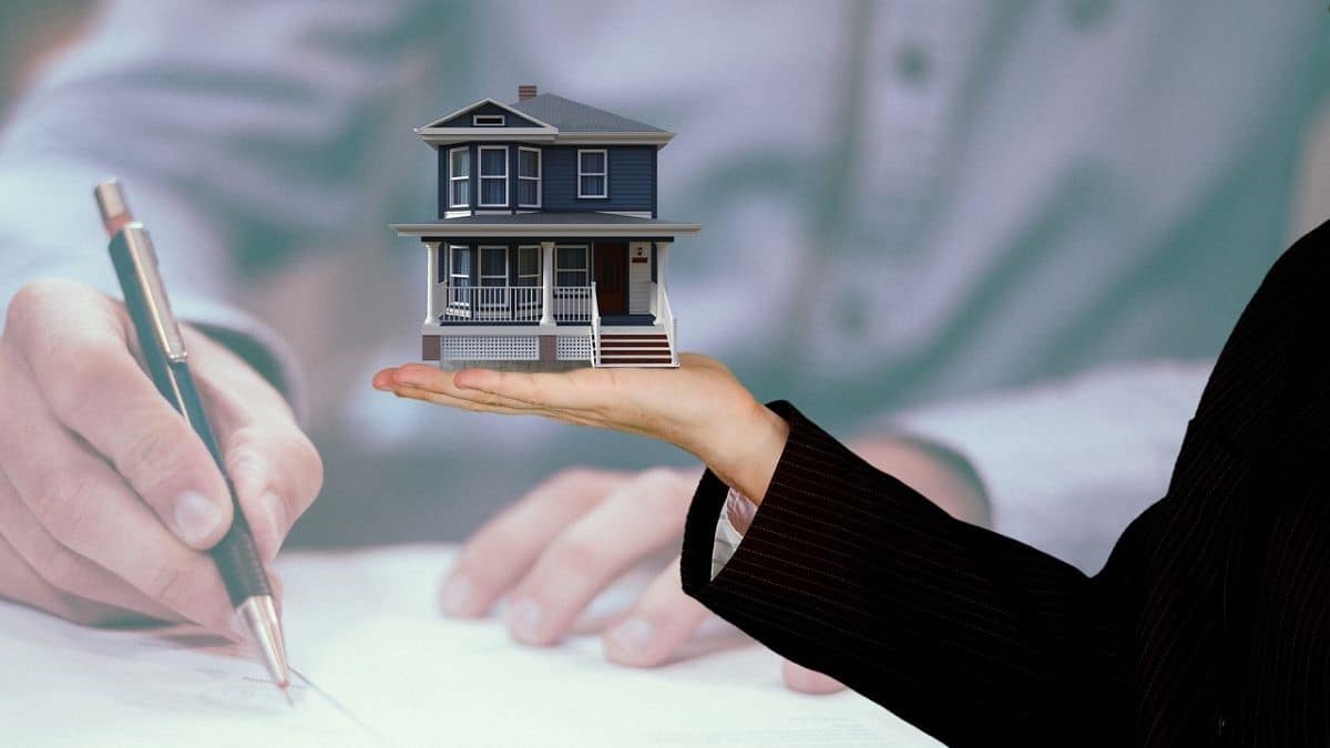 successful real estate agents represented by a businesswoman's hand holding up a model of a new home against a muted background of a man's hands signing legal documents