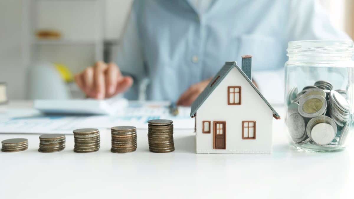 mortgage lending represented by stacks of coins and a jar or coins next to a small model house, with a businessman using a calculator in the background