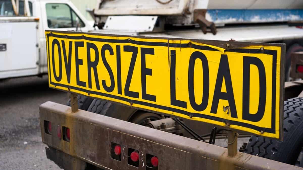 heavy haul loads represented by a black-on-yellow oversize load sign installed on the back of big rig semi truck tractor with heavy metal bumper for safety standing on a parking lot awaiting an oversized cargo load